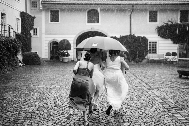 Braut und Brautjungfer laufen mit Regenschirm über einen nassen Innenhof
